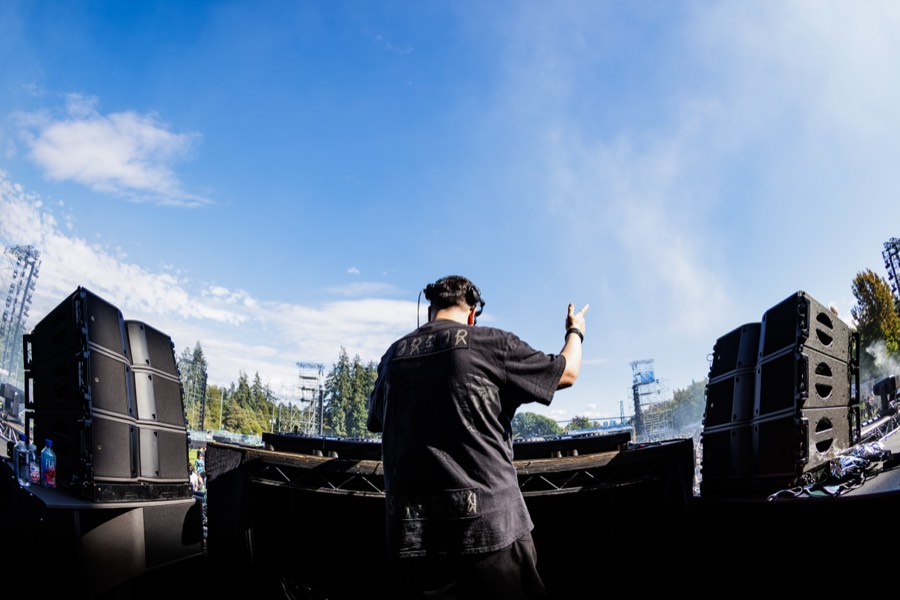 A DJ raises a hand to the crowd from behind the decks under a clear afternoon sky