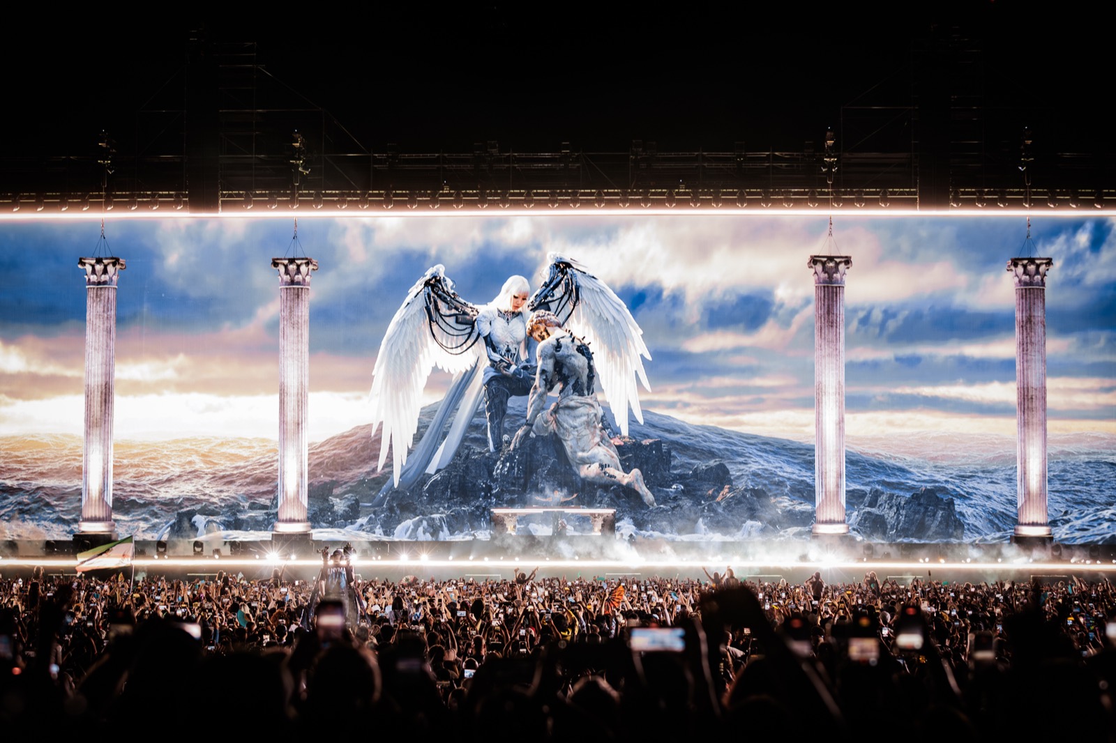The ÆDEN stage at full scale — a winged figure rising over stormy seas across the LED wall, framed by illuminated columns, above a crowd of raised phones